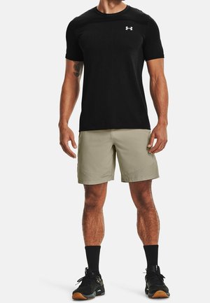 T-shirt de sport noir à manches courtes avec logo Under Armour, associé à un short beige clair avec un ourlet texturé et des chaussures de sport noires.
