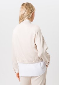 Beige bomberjack met een gladde, lichte stof. Heeft een staande kraag en een elastische zoom, gedragen over een wit shirt.