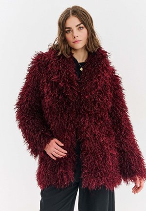 Kobieta ubrana w puszysty, głęboko czerwony płaszcz na czarnym ubraniu, stojąca z jedną ręką na biodrze na tle jednolitej, białej ściany.