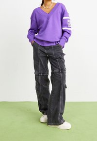Purple v-neck sweatshirt with white accents paired with black cargo pants featuring pockets and contrasting stitching, worn with white sneakers.