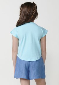 Svetlo modra majica s kratkimi rokavi in zaobljenim robom, v kombinaciji z svetlim denim kratkimi hlačami, prikazana s hrbtne strani.