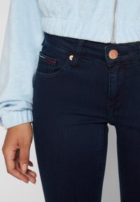Jeans de mezclilla azul oscuro con una forma ajustada. Presenta un botón de cobre y un pequeño bolsillo con detalles de rayas rojas y blancas.