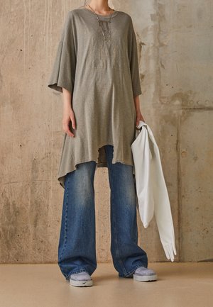 Persona con túnica gris holgada, vaqueros azules de pierna ancha, zapatillas grises, sosteniendo una prenda blanca frente a un fondo de pared de hormigón texturizado.