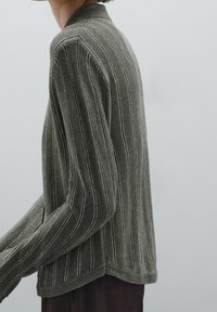 Person wearing a long-sleeve, ribbed, grayish-green sweater with vertical white stripes, standing against a plain light background.