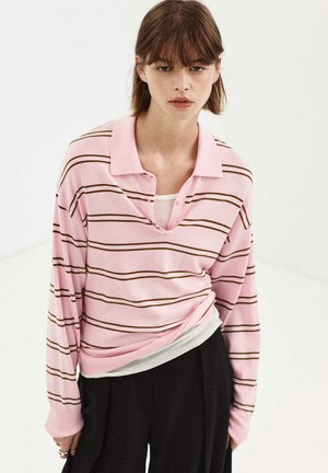 Mujer joven con cabello castaño que lleva una camisa polo de manga larga a rayas en rosa y marrón y pantalones negros sobre un fondo liso claro.