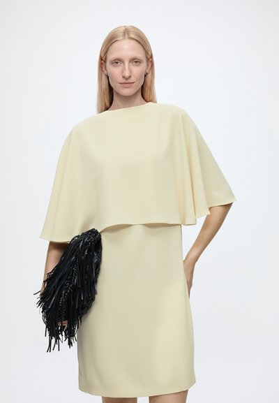 Femme aux cheveux blonds et raides porte une robe ample couleur crème et tient un sac à main noir à franges devant un fond blanc uni.