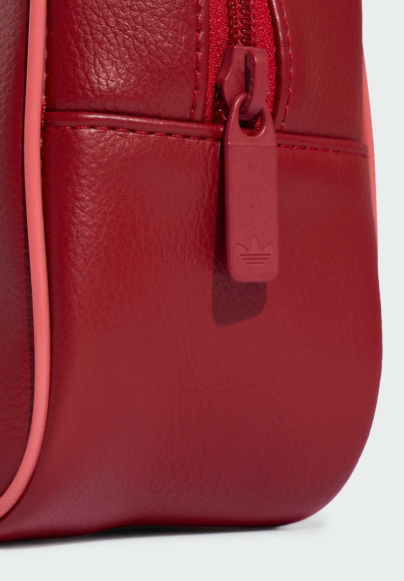 Primo piano dell'angolo di una borsa in pelle rossa con cerniera rossa abbinata e linguetta con logo Adidas in rilievo.