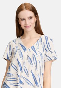 Kurzärmlige Bluse mit V-Ausschnitt, auf cremigem Hintergrund mit blauen Fischmustern. Hergestellt aus leichtem Stoff.