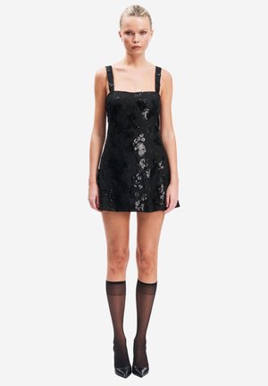 Frau trägt ein ärmelloses schwarzes Minikleid mit Blumenmuster, transparente schwarze knielange Socken und schwarze spitze High Heels, steht vor weißem Hintergrund.
