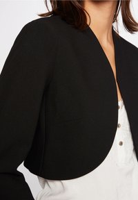 Woman wearing a cropped black jacket with curved hem over a white lace-trimmed blouse with buttons, shown from neck to waist.