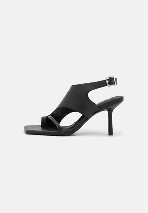 Sort læderhøjhælet sandal med åben tå, udskårne sider, tynd rem over tæerne og ankelrem med spænde på hvid baggrund.