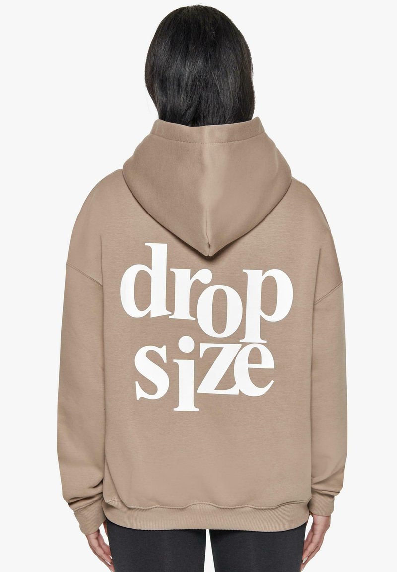 Beige Kapuzenpullover mit weißem Text "drop size" auf dem Rücken, ausgestattet mit einer geräumigen Kapuze und gerippten Bündchen, hergestellt aus einem weichen, strukturierten Stoff.