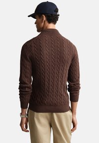 Bruine kabelgebreide trui met een geribde kraag en manchetten, met een gestructureerd patroon. Gedragen met kaki broek. Achteraanzicht weergegeven.