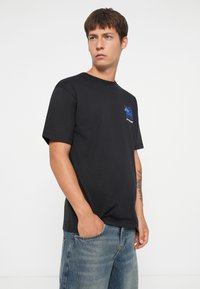 Zwarte katoenen t-shirt met korte mouwen, met een blauw grafisch ontwerp op de linkerkant van de borst. Draagt met lichtblauwe jeans.