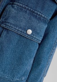 Gros plan sur un tissu en denim bleu avec une poche à rabat fermée par un bouton-pression argenté gravé "Bershka."
