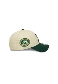 Casquette de baseball crème avec une visière verte. Dotée d'un patch circulaire brodé avec un ours et un texte, ainsi qu'un logo sur le côté cousu en vert.