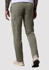 Pantalones cargo verde oliva con bolsillos laterales, corte ajustado y textura sutil; llevados con zapatillas de colores claros, vistos desde atrás.