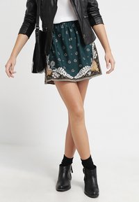 Groene rok met een patroon van bloemen en gouden accenten, met een uitlopende vorm en een geplooid tailleband, gecombineerd met zwarte enkellaarsjes.