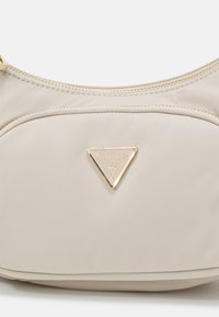 Sac à main en tissu beige avec une fermeture éclair dorée et un logo triangulaire doré Guess sur la poche avant centrale.