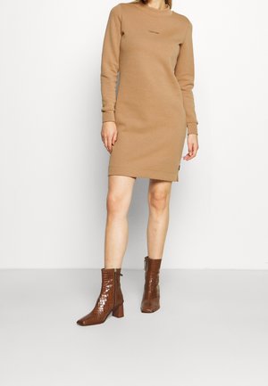 Femme portant une robe Calvin Klein camel à manches longues, associée à des bottines marron brillantes à talon bloc.