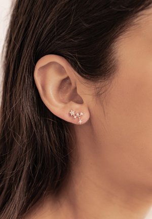 Ear with dark hair wearing small rose gold star-shaped earrings connected by a delicate chain.