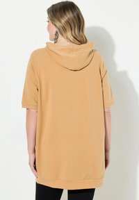 Tunique beige courte avec capuche et manches courtes, offrant une coupe décontractée et un ourlet qui descend au-delà des hanches. Texture lisse et douce.
