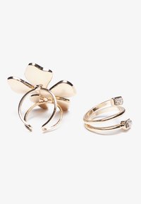 Bague florale en ton d'or avec une surface lisse et polie et des pétales dorés ; à côté, une bague double minimaliste avec deux accents carrés.