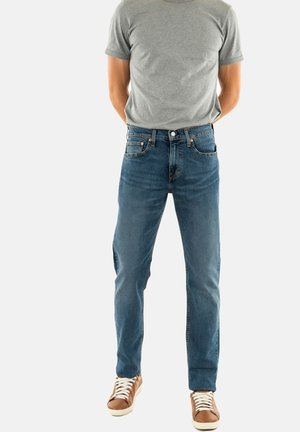 Camiseta de manga corta gris claro combinada con vaqueros rectos de color azul. Los vaqueros presentan un diseño clásico de cinco bolsillos y un ligero desgaste. Zapatillas marrones.