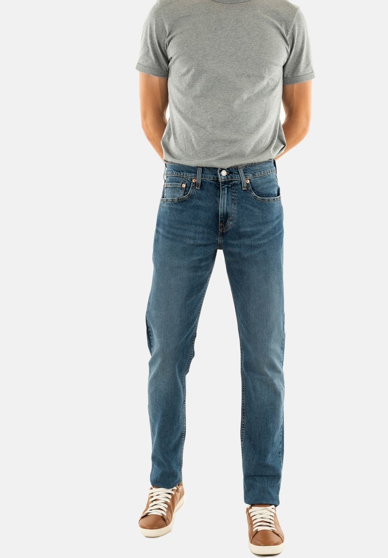 Camiseta de manga corta gris claro combinada con vaqueros rectos de color azul. Los vaqueros presentan un diseño clásico de cinco bolsillos y un ligero desgaste. Zapatillas marrones.