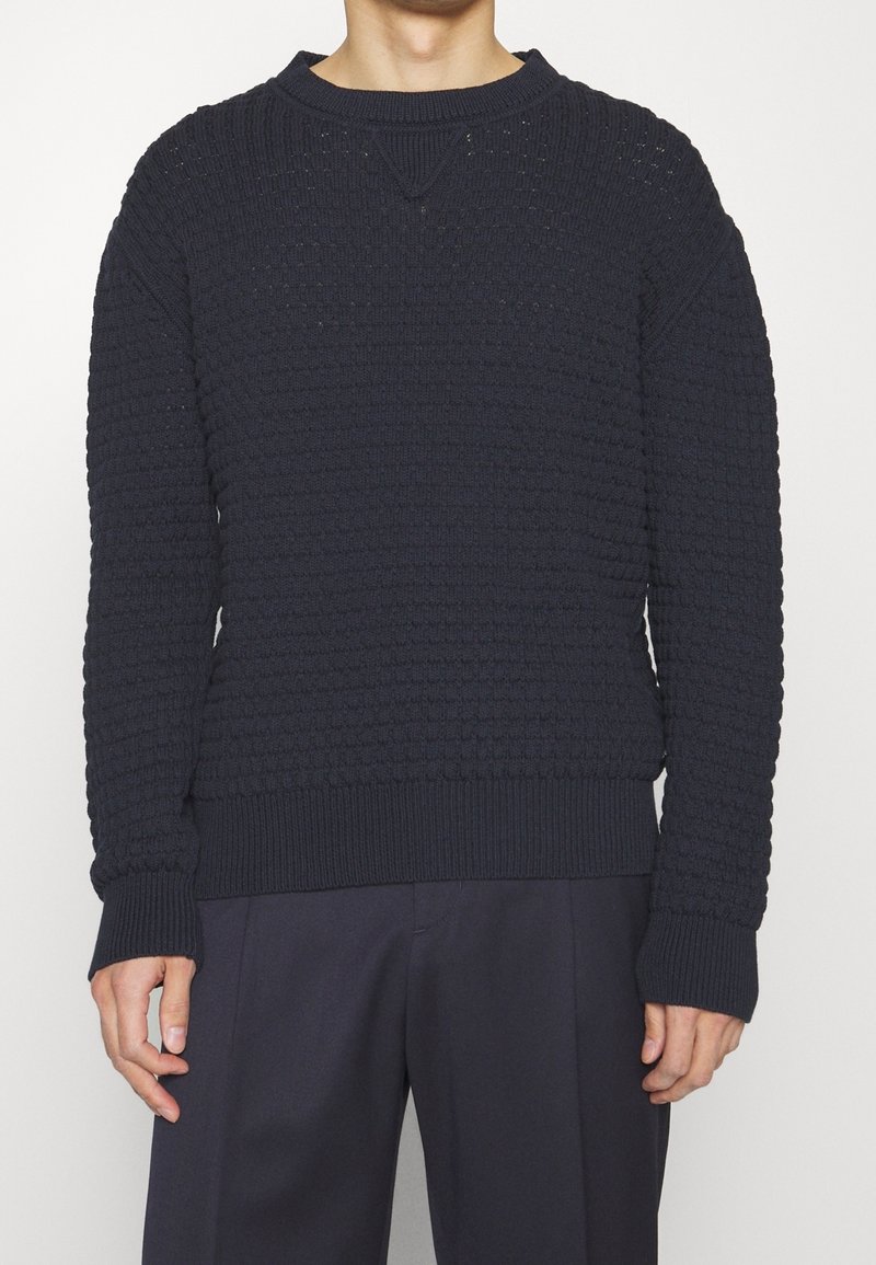 Homme portant un pull en maille texturée bleu marine foncé avec poignets et ourlet côtelés, associé à un pantalon foncé sur un fond uni.