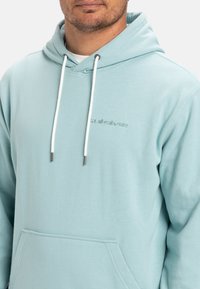 Light blue hoodie made of soft fabric, featuring a kangaroo pocket, white drawstrings with metal tips, and a subtle logo on the chest.