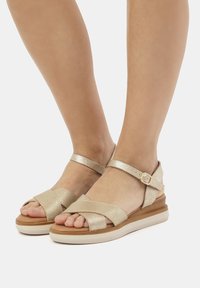 Goldene metallische Sandalen mit überkreuztem Vorderdesign und Knöchelriemen. Dicke Sohle mit braunem Grund und hellfarbigem Boden.