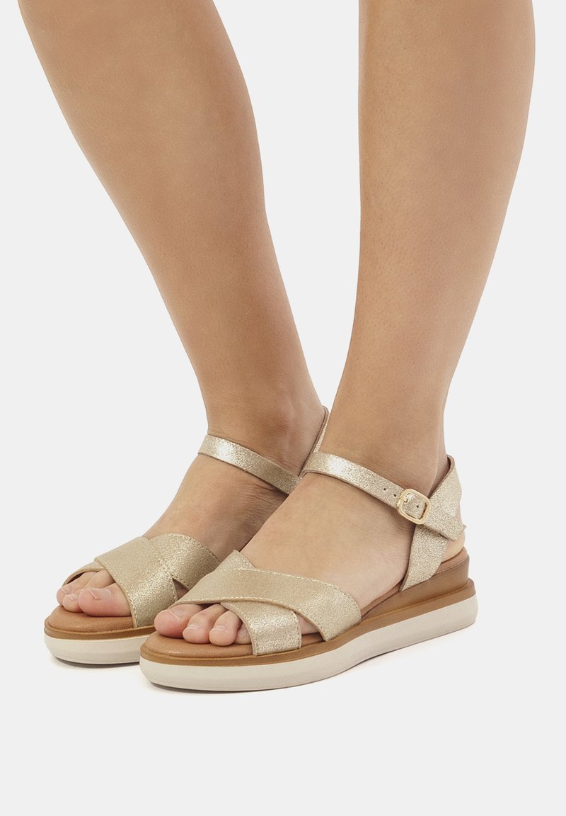 Goldene metallische Sandalen mit überkreuztem Vorderdesign und Knöchelriemen. Dicke Sohle mit braunem Grund und hellfarbigem Boden.