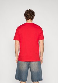 Červené bavlněné tričko s krátkými rukávy a kulatým výstřihem, zobrazené zezadu. Doplněno světle modrými džínovými šortkami s kapsami.