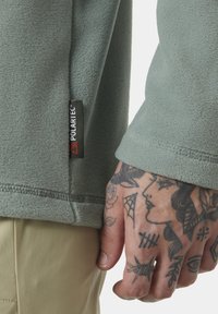 Fleece bunda z jemného, světlého zeleného materiálu; na boku je štítek Polartec, s detailním záběrem potetované ruky, která drží rukáv.