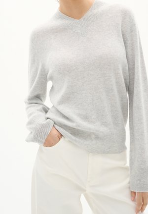 Mujer con suéter gris claro de cuello en V y mangas largas metido dentro de pantalones blanco hueso, con una mano en el bolsillo, de pie contra un fondo blanco.