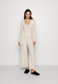 Beige lounge set featuring a soft, v-neck top and wide-leg pants with an elastic waistband. Paired with black slip-on slippers.