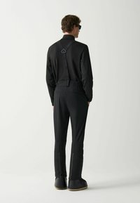 Uomo che indossa pantaloni neri attillati con bretelle, maglia nera a maniche lunghe, occhiali da sole neri e stivali neri, in piedi contro uno sfondo bianco.