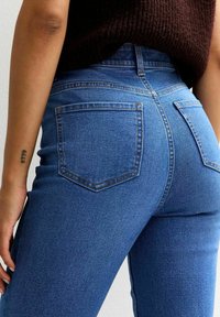 Persona con jeans blu a vita alta e un maglione lavorato a maglia marrone scuro, vista di schiena e di lato con un piccolo tatuaggio sul braccio sinistro.