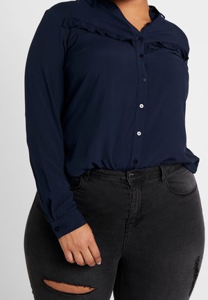 Nainen pukeutunut laivastonsiniseen, pitkähihainen napitettavaan paitaan, jossa on röyhelöt, ja mustiin revittyihin farkkuihin, yksinkertaisen taustan edessä.