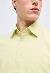 Homme portant une chemise boutonnée jaune clair avec un col pointu et un logo brodé "BOSS" sur la poitrine.