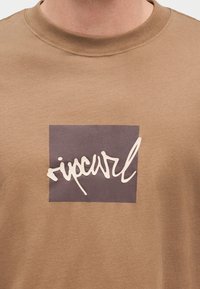 Καφέ μπλουζάκι με σκοτεινό τετράγωνο και άσπρο καλλιγραφικό κείμενο "ripcurl" τυπωμένο στο στήθος.