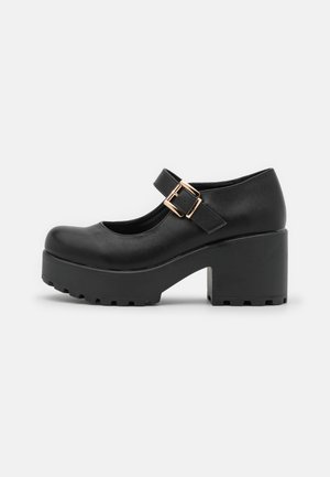 Schwarze Mary Jane Schuhe mit rundem Zehenbereich, einer seitlichen Schnalle und einer grob strukturierten, klobigen Sohle. Der Absatz ist etwa 10 cm hoch.