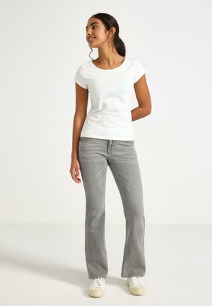 Giovane donna in piedi, indossa una maglietta bianca a maniche corte, jeans grigi e scarpe da ginnastica bianche, guarda di lato con una mano dietro la schiena.