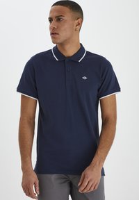 Mørkeblå polo-skjorte med en to-knaps placket, hvit krage og mansjettkanter. Har en liten logo på venstre bryst. Laget av bomull.