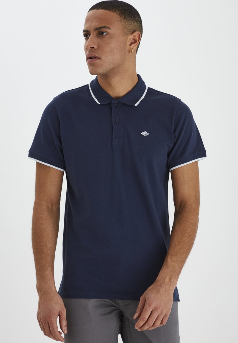 Mørkeblå polo-skjorte med en to-knaps placket, hvit krage og mansjettkanter. Har en liten logo på venstre bryst. Laget av bomull.