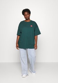 Groene oversized katoenen t-shirt met een ronde halslijn en korte mouwen, voorzien van een kleine roze bloemenborduursel op de linker borst.