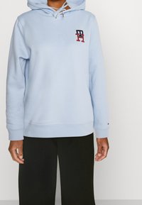 Jasnoniebieska bluza z kapturem z miękkiego materiału z logo na przodzie, kapturem z regulowanym sznurkiem i ściągaczami na rękawach. Zestawiona z czarnymi szerokimi spodniami.