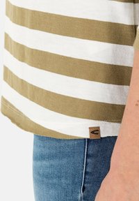 Chemise à rayures horizontales beige et blanc associée à un jean en denim bleu, avec une petite étiquette marron visible sur l'ourlet de la chemise.