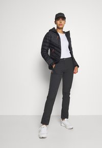 Zwarte pufferjack met rode accenten, witte t-shirt en zwarte broek. Schoenen zijn witte sportieve schoenen. Model staat zelfverzekerd tegen een neutrale achtergrond.